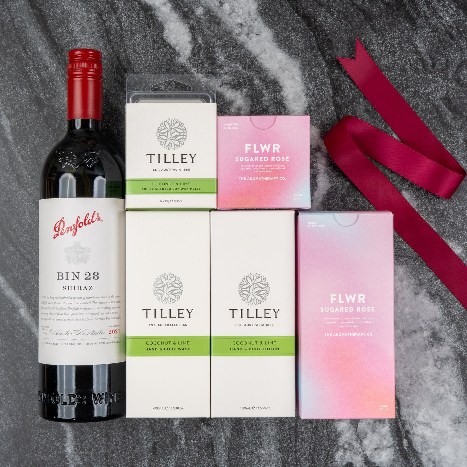 The Hamper Boutique Co's Penfolds Spa and Indulgence Hamper features Penfolds Bin 28 Shiraz wine, Tilley Coconut & Lime soap, hand & body lotion, a candle, and two FLWR Sugared Rose candles arranged on gray marble with a pink ribbon.