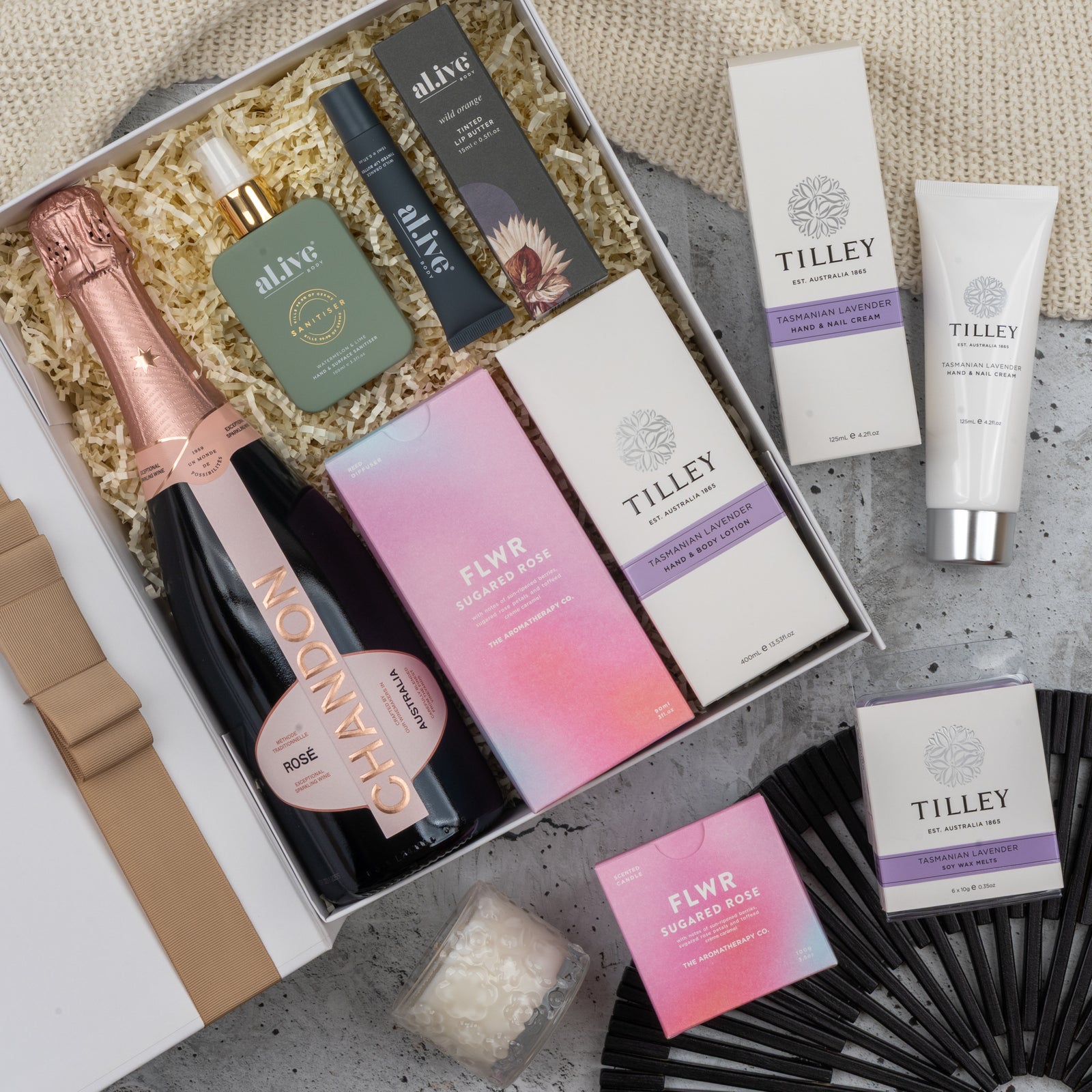 The Chandon Rosé Pamper Hamper by The Hamper Boutique Co features a bottle of Chandon rosé, skincare items, candles, soaps, and a black hand fan arranged on shredded paper with a beige cloth backdrop.