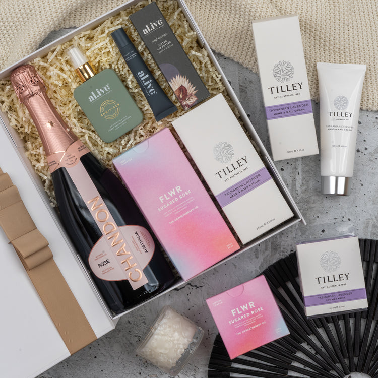 The Chandon Rosé Pamper Hamper by The Hamper Boutique Co features a bottle of Chandon rosé, skincare items, candles, soaps, and a black hand fan arranged on shredded paper with a beige cloth backdrop.
