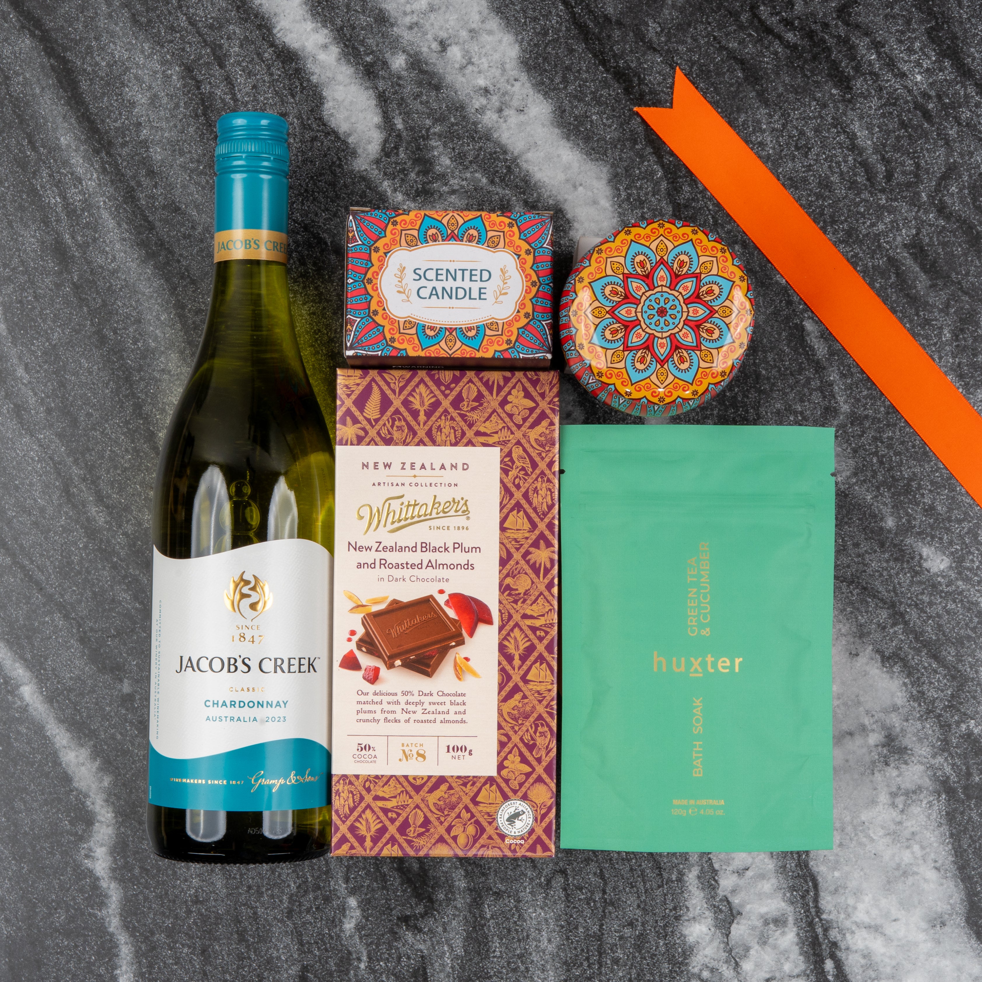 The Hamper Boutique Co’s Wine and Candlelight Hamper, featuring wine, a candle, chocolate, soap pouch, and a decorative tin, is displayed on gray marble with an orange ribbon.