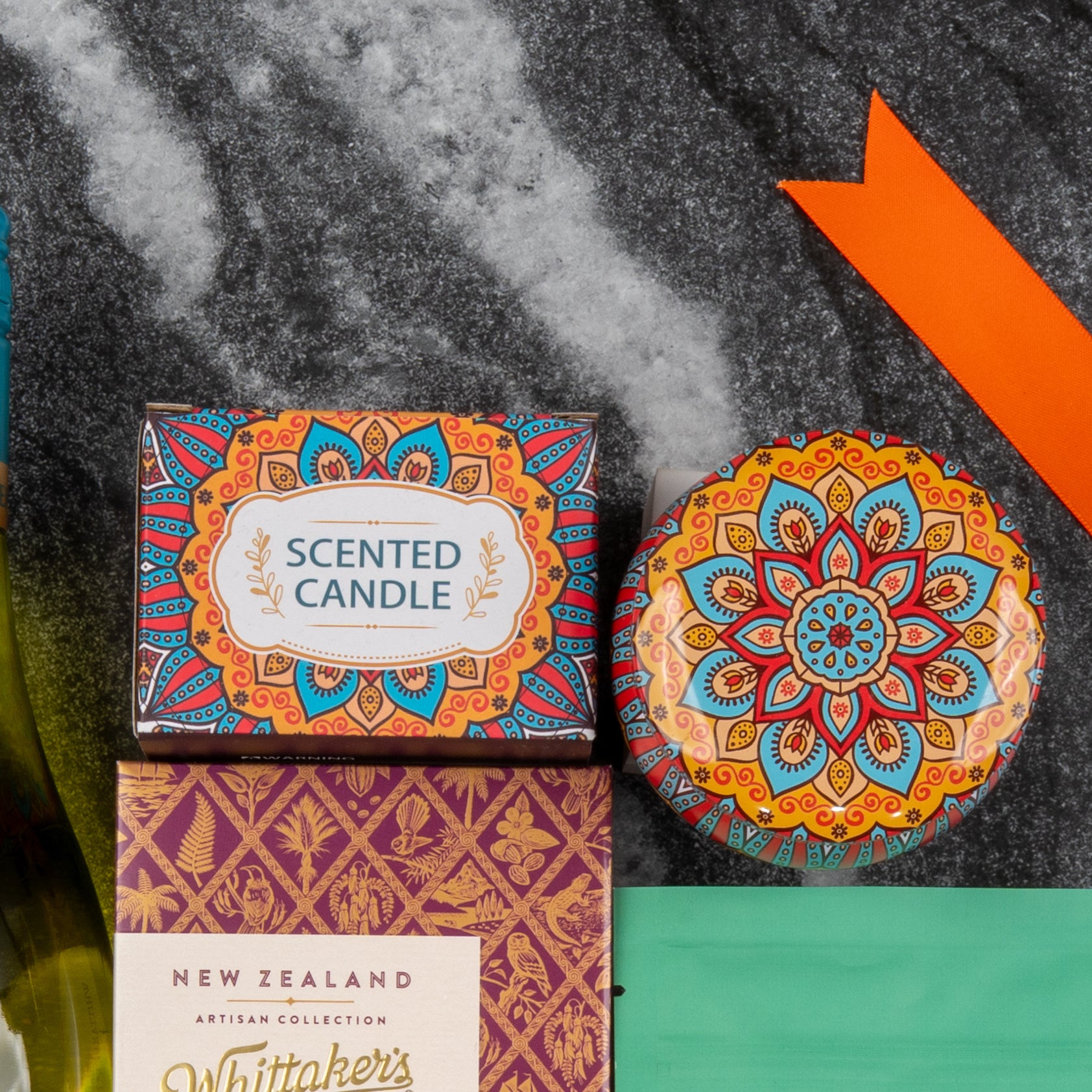 The Hamper Boutique Co's Wine and Candlelight Hamper features a decorative scented candle in a mandala tin, set on a dark marbled surface beside a gold-and-purple Whittakers chocolate box and an orange ribbon.