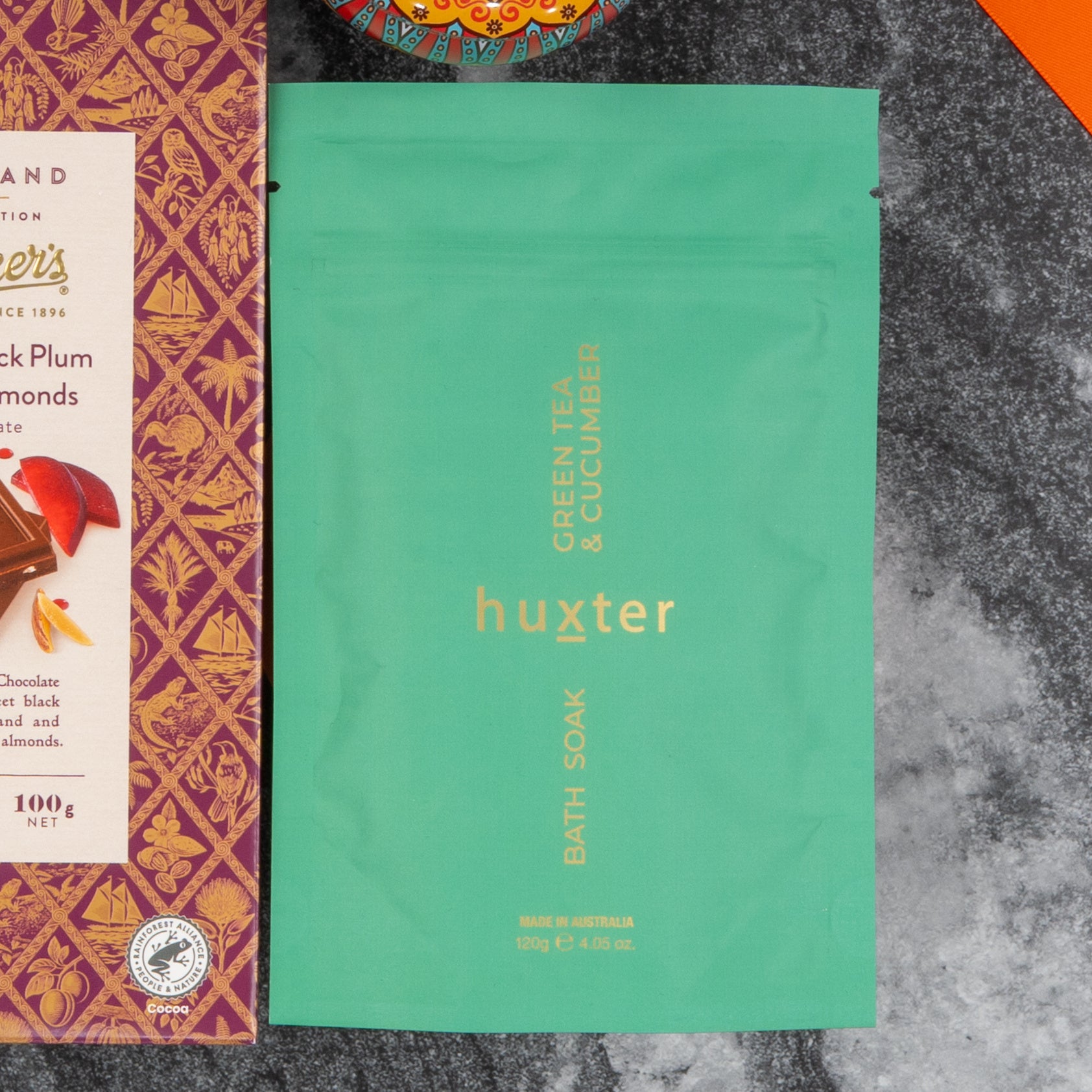 A green Huxter Green Tea & Cucumber bath soak pouch from The Hamper Boutique Co’s Wine and Candlelight Hamper sits by a patterned chocolate box on a dark marbled surface, with part of a colorful bowl at the top of the image.