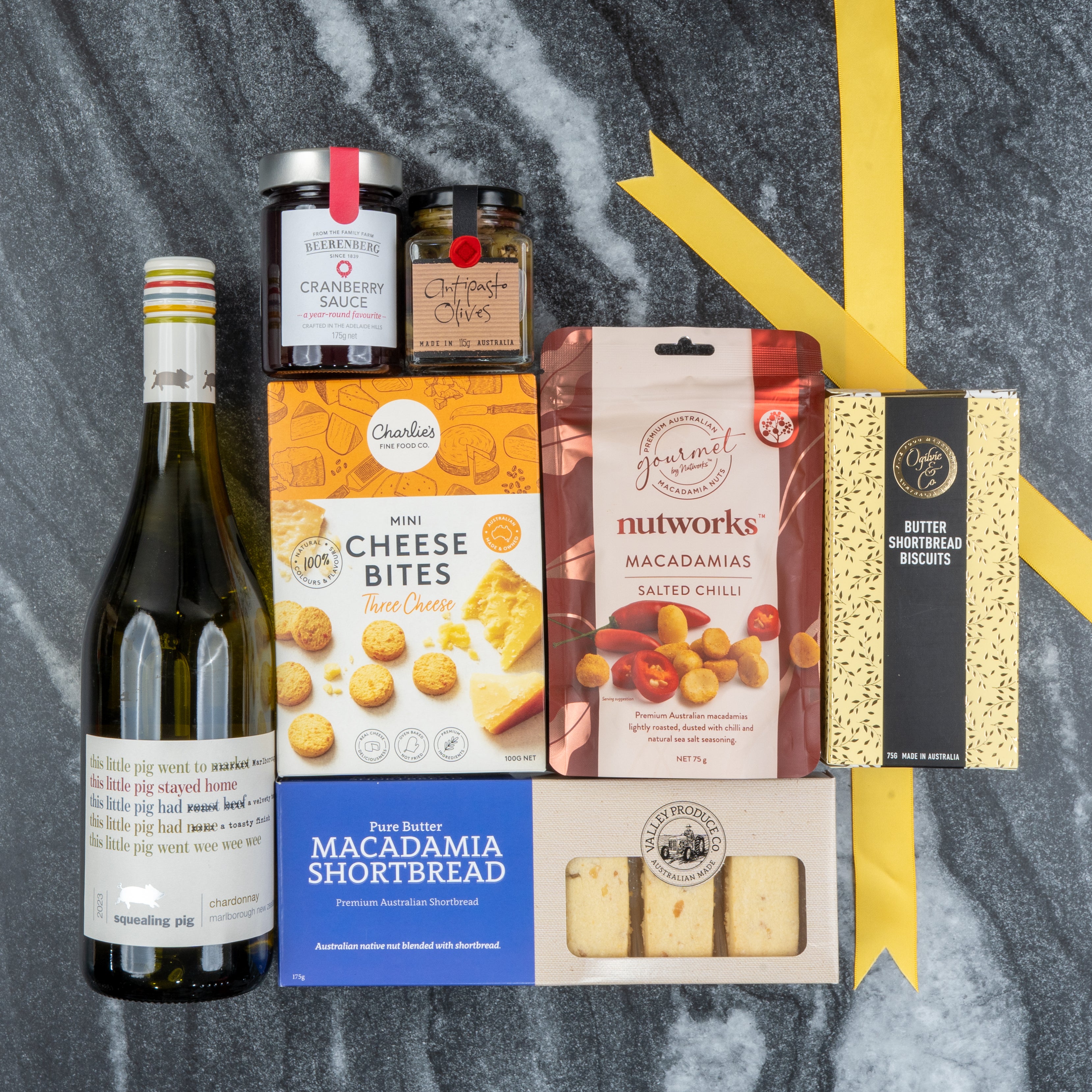 The Hamper Boutique Co’s White Wine Gourmet Hamper features white wine, cranberry sauce, fig paste, olives, cheese bites, salted chili macadamias, macadamia shortbread, and butter biscuits on dark marble with a yellow ribbon.