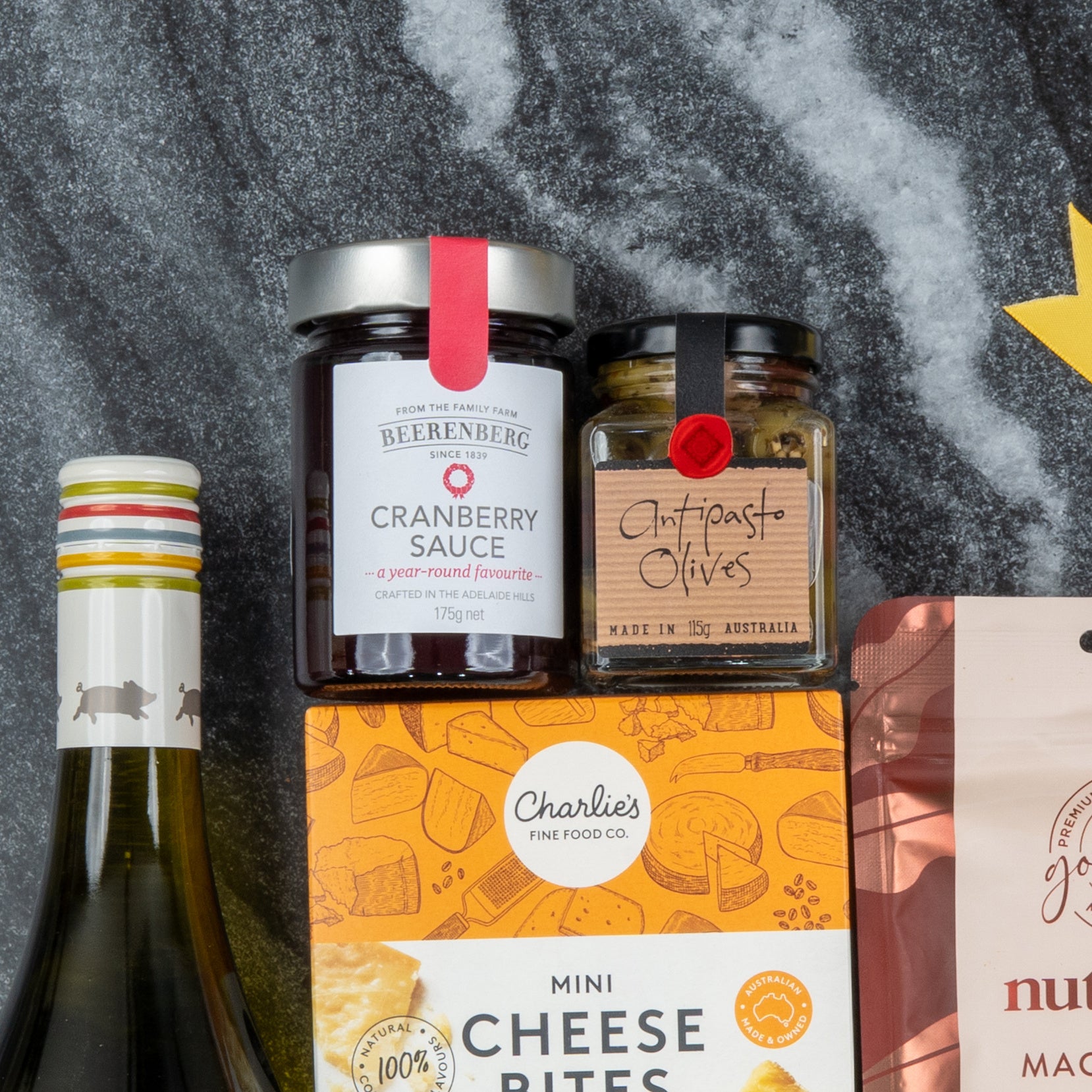 The Hamper Boutique Co's White Wine Gourmet Hamper, featuring white wine, cranberry sauce, antipasto olives, and mini cheese bites, is arranged on a gray surface with part of a pink snack package visible on the right.