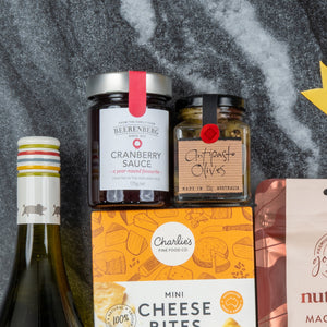 The Hamper Boutique Co's White Wine Gourmet Hamper, featuring white wine, cranberry sauce, antipasto olives, and mini cheese bites, is arranged on a gray surface with part of a pink snack package visible on the right.