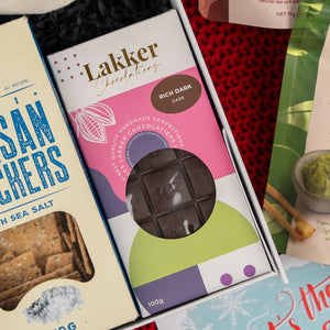A box of Cheers & Tonic by The Hamper Boutique Co is placed next to a selection of other food items and sea salt crackers on a red textured surface, with its contents visible through a clear window.