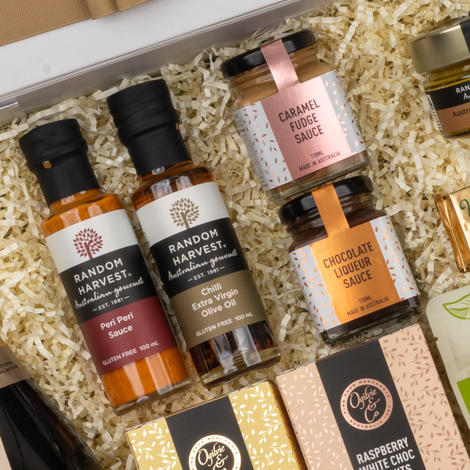The Hamper Boutique Co Chandon Gourmet Celebration Hamper features gourmet treats on straw-like packing, including Peri Peri Sauce, Chilli Extra Virgin Olive Oil, Caramel Fudge Sauce, and Chocolate Liqueur Sauce in glass bottles and jars.