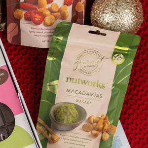 The Hamper Boutique Co's Cheers & Tonic features a gold glitter ornament, a green Nutworks Macadamias Wasabi bag, assorted chocolates, and a red knitted backdrop for a festive touch.