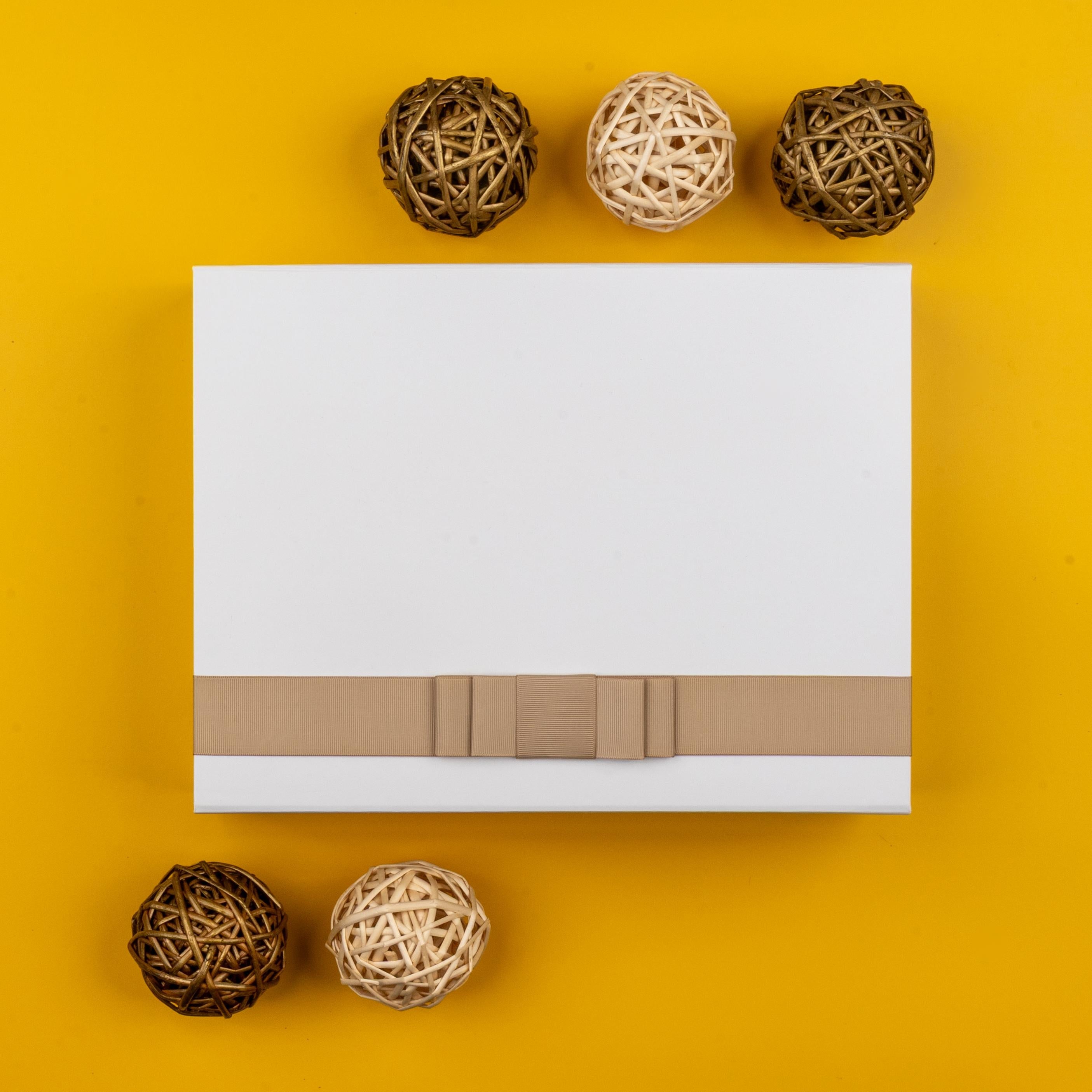 The Hamper Boutique Co’s Elegance and Self-Care Gift Hamper is presented in a white gift box with a beige ribbon, centered on a yellow background and encircled by six light and dark brown wicker balls.