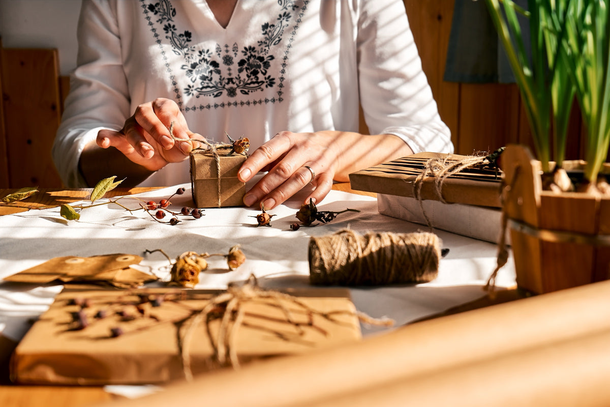 Hands tying twine around small gifts wrapped in brown paper, with dried flowers on the table