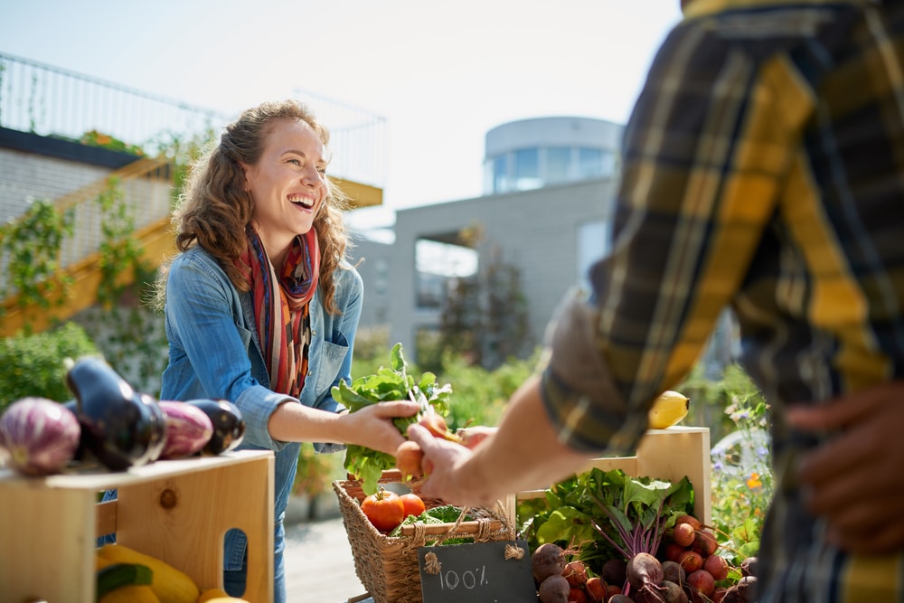 A smiling stallholder hands fresh produce to a customer at an outdoor market