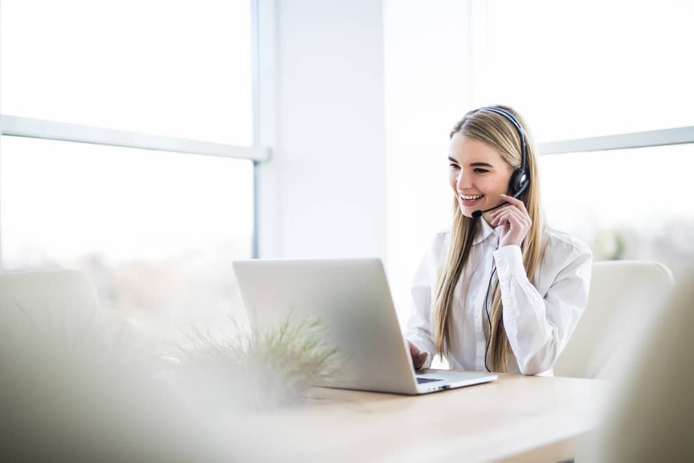 A smiling woman with a headset working at a laptop in a bright office